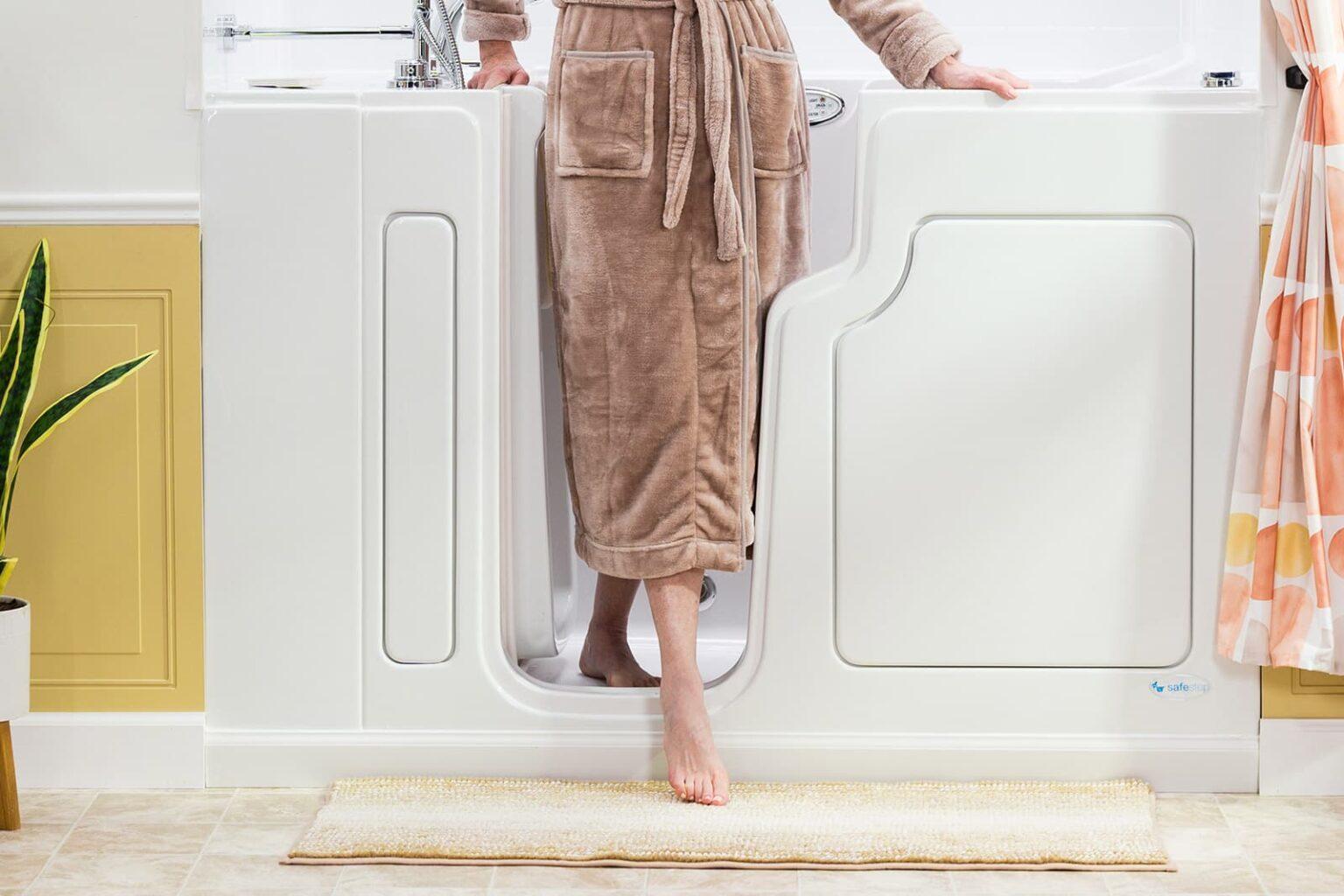 SafeStep walk-in tub featuring ultra-low threshold design with person in beige robe safely stepping into white tub with easy-entry door in modern bathroom